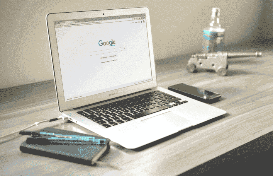 Como anunciar no Google Ads: em uma mesa, estão dispostos um caderno com canetas e um computador. A tela do computador está ligada na página de busca do Google, dando para ver a logo colorida em vermelho, amarelo, verde e azul da empresa.