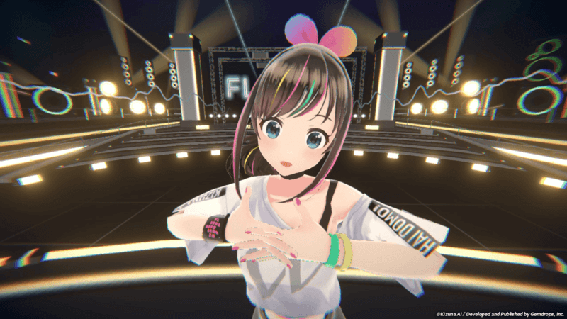 VTuber 11 Imagem da personagem virtual Kizuna AI em um palco iluminado com luzes e elementos de show ao fundo. Ela está no centro, olhando para a câmera, com expressão levemente surpresa, vestindo blusa branca de ombro caído com a palavra “HALO DOMO!” na manga, pulseiras coloridas e laço rosa e azul na cabeça.